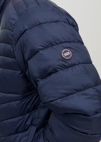 Tumedat sinine puffer-jakk neljaõmmelise disainiga. Selles on ümmargune logotäkk varrukal ja sile, läikiv tekstuur.