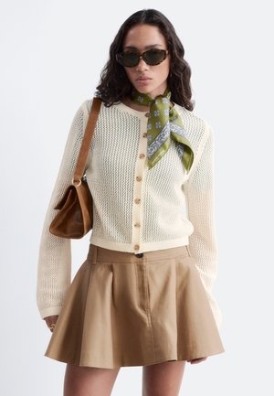 Femme portant des lunettes de soleil marron, un foulard vert à motifs autour du cou, un cardigan en maille crème, une jupe plissée marron et un sac à bandoulière en cuir marron.