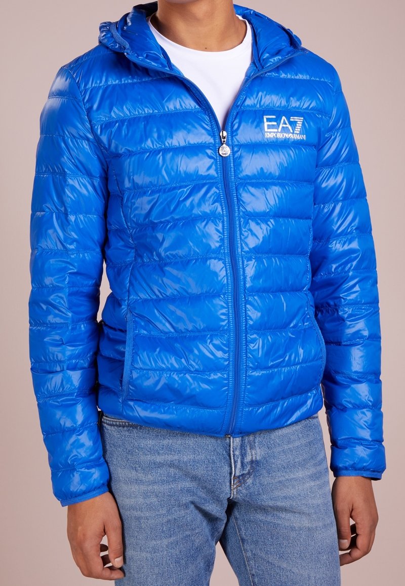 Niebieska kurtka puchowa z błyszczącym wykończeniem, z zamkiem błyskawicznym, kapturem i ukośnymi wzorami przeszyć. Posiada dwie boczne kieszenie oraz logo na piersi.