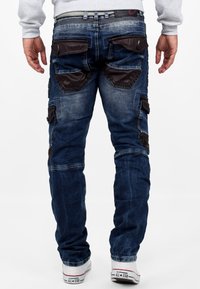 Jeans en denim bleu avec des accents en cuir noir sur les poches, design slim-fit, couture visible et ourlets retroussés. Associés à des baskets blanches.