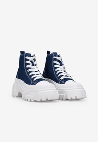 Zapatillas altas de denim azul con cordones blancos y una suela gruesa. Las características incluyen dos lengüetas y punteras de goma blanca.
