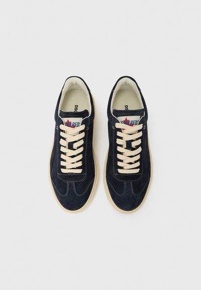 Tengerészkék színű, velúr sportcipők krémszínű fűzőkkel, lekerekített orral, kontrasztos varrással és márkajelzéssel a nyelvnél. Lapos gumitalp.