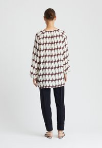 Lange mouwen, crèmekleurige blouse met een marron geometrisch patroon, gecombineerd met donkere broek. De stof lijkt lichtgewicht met geplooide details bij de halslijn.