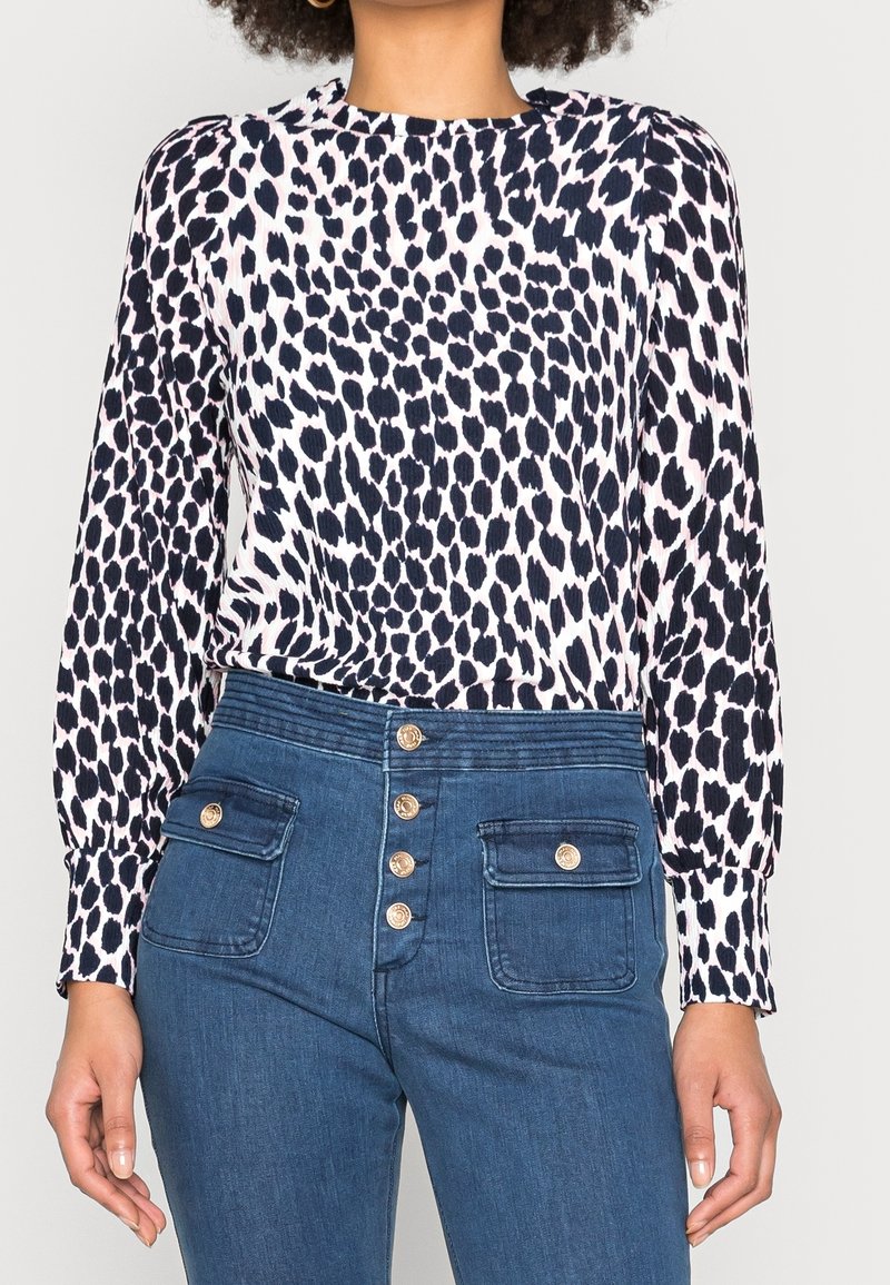 Femme portant une blouse à manches longues imprimé léopard noir et blanc rentrée dans un jean bleu taille haute avec des boutons dorés et des poches à rabat devant.
