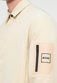 Μπεζ πουκάμισο με μακριά μανίκια, γιακά και τσέπη με φερμουάρ στο πάνω μέρος του μπράτσου, με κεντημένη ετικέτα με το λογότυπο BOSS.
