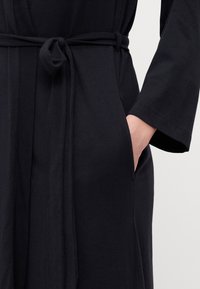Robe en coton noir avec des manches longues, une taille nouée et une poche latérale. Texture lisse et design simple.