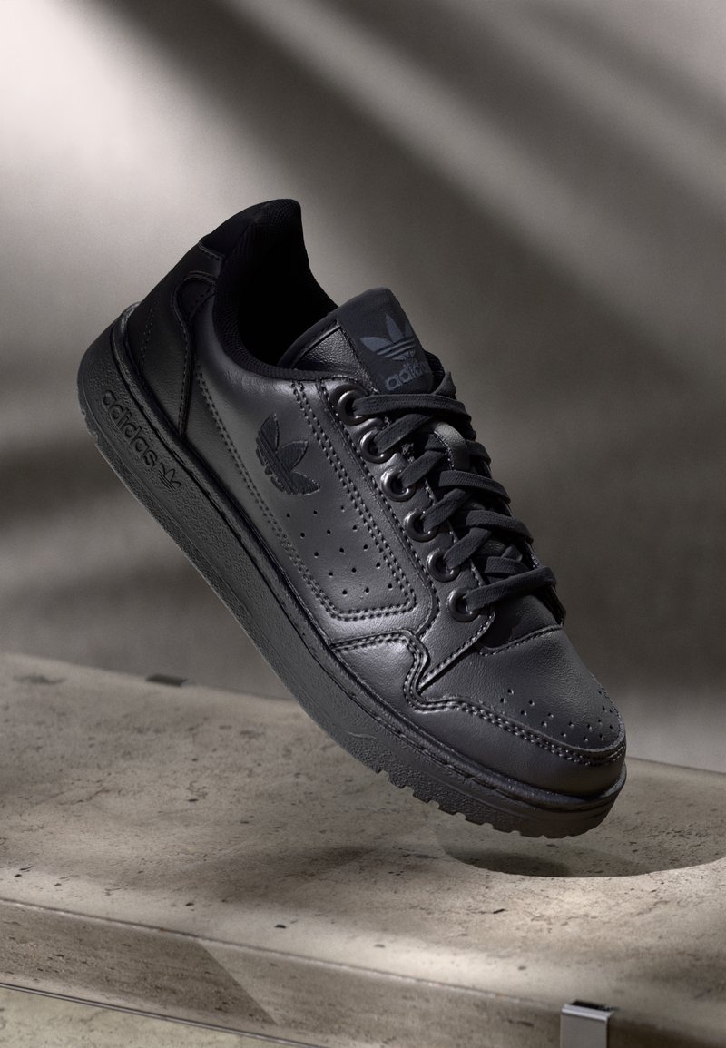 Černé kožené tenisky Adidas s logem na boku a jazyku, černá šňůrka, odpočívající na betonovém povrchu s jemným stínem v pozadí.