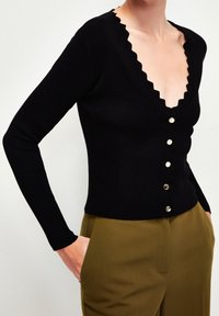 Donna che indossa un cardigan nero a maniche lunghe con bottoni e bordi ondulati, abbinato a pantaloni verdi oliva a vita alta, con le mani in tasca.