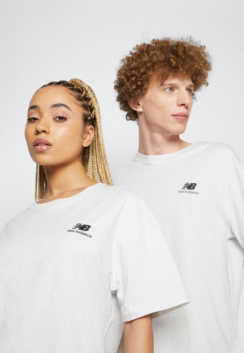 Due persone indossano t-shirt a maniche corte di un grigio chiaro con un logo nero "New Balance" sul petto. Il materiale sembra morbido e informale.