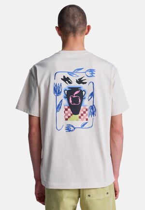 Personne portant un t-shirt blanc avec un vase noir, une pomme rose, des fleurs bleues, deux oiseaux noirs et un motif à damier rouge dans le dos.