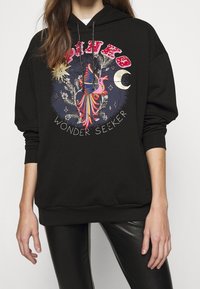 Svart hoodie med en stor färgglad grafik som har en stiliserad fågel och texten "WONDER SEEKER", accentuerad med stjärnor och en halvmåne.