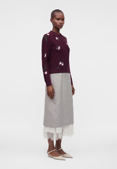 Femme debout de profil, portant un pull bordeaux à fleurs, une jupe grise avec ourlet en dentelle et des ballerines crème à bouts pointus, sur un fond uni.