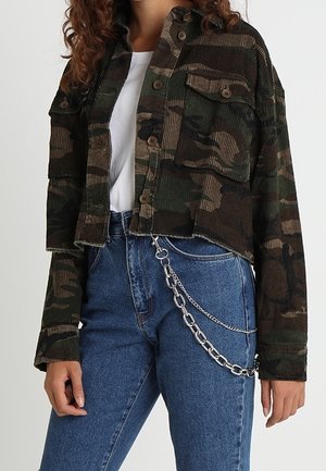 Vrouw die een cropped camouflagejack draagt over een wit shirt, met een hooggetailleerde blauwe jeans en een zilveren kettingaccessoire aan de rechterkant.
