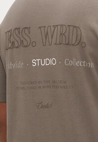 Pilka medvilninė marškinėlių su reljefiniu ir atspausdintu tekstu. Ryškūs elementai – „ESS. WRD.“ ir „STUDIO“ skirtingų spalvų.