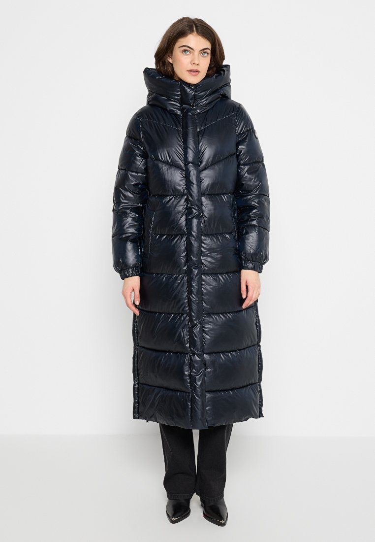 Manteau matelassé long, bleu marine foncé avec un col haut et une finition brillante. Présente des coutures verticales, une fermeture éclair et des poches latérales.