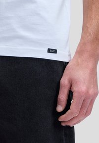 Közelkép egy kézről, amely fekete farmernadrág mellett, egy fehér póló alsó szélén, kis fekete Lee márkajelzéssel pihen.