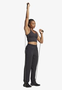 Schwarzes Sportoberteil und lockere schwarze Sweatpants, hält Widerstandsbänder während sie steht. Glatte Texturen mit einer figurbetonten Silhouette.