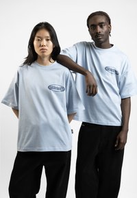 Light blue oversized t-shirts with a small dark logo, worn with black pants. Soft cotton fabric and relaxed fit.