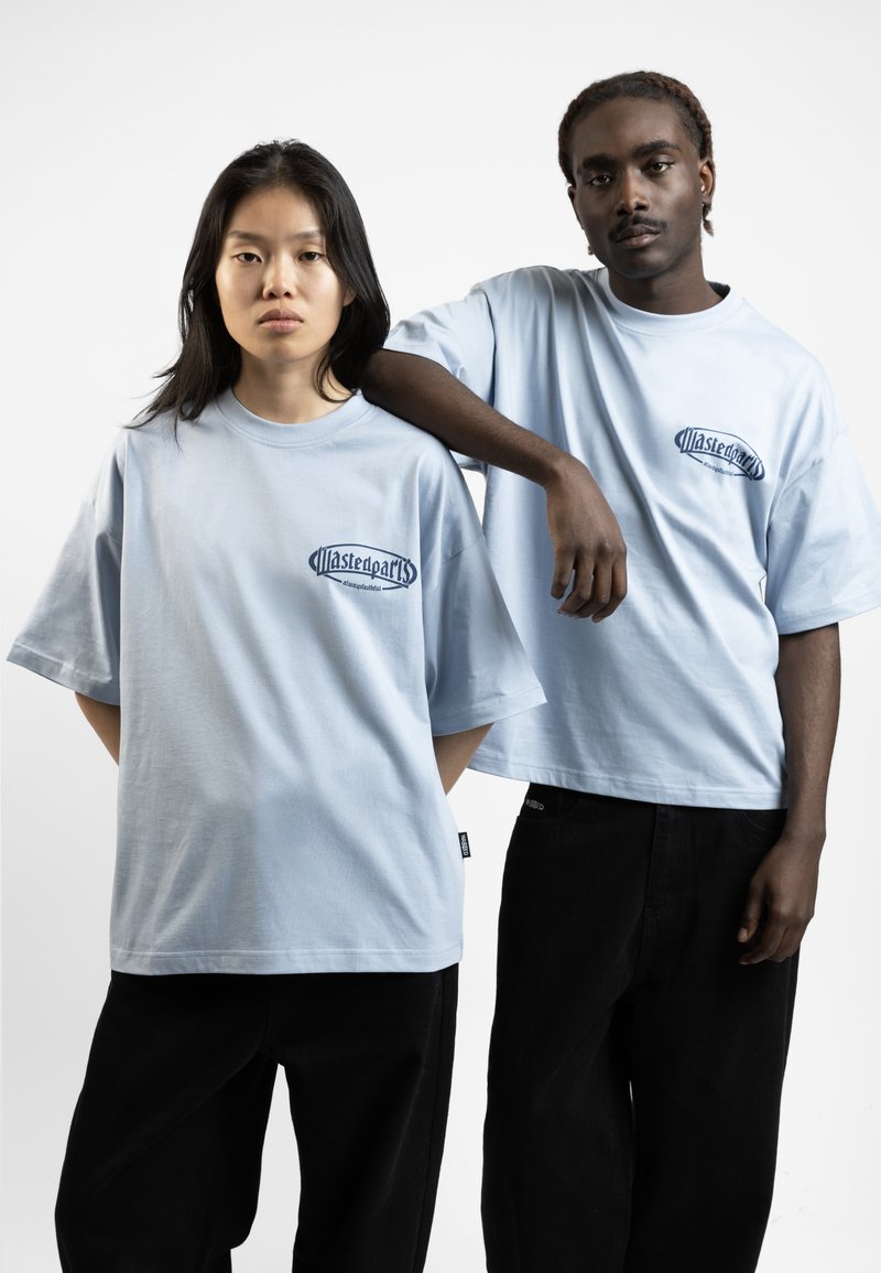 Light blue oversized t-shirts with a small dark logo, worn with black pants. Soft cotton fabric and relaxed fit.