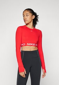Crvena dugih rukava skraćena majica s okruglim izrezom i bijelim logotipom "NIKE PRO" na pojasu. Uparena s crnim tajicama visokog struka koje imaju kontrastne šavove.