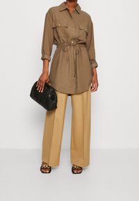 Halenka s dlouhým rukávem, na knoflíky, hnědé barvy s předními kapsami a stahovacími šňůrkami v pase, v kombinaci s béžovými kalhotami širokého střihu a černou texturovanou taškou.
