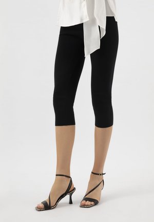 Femme portant des leggings noirs courts et des sandales noires à talons hauts avec des brides à la cheville, sur un fond blanc uni.