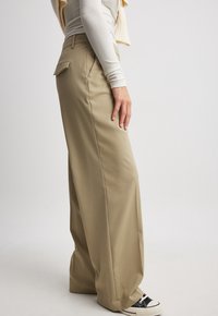 Person som bär beige vida byxor, krämfärgad långärmad topp och svart-vita sneakers, stående på sidan mot en enfärgad bakgrund.