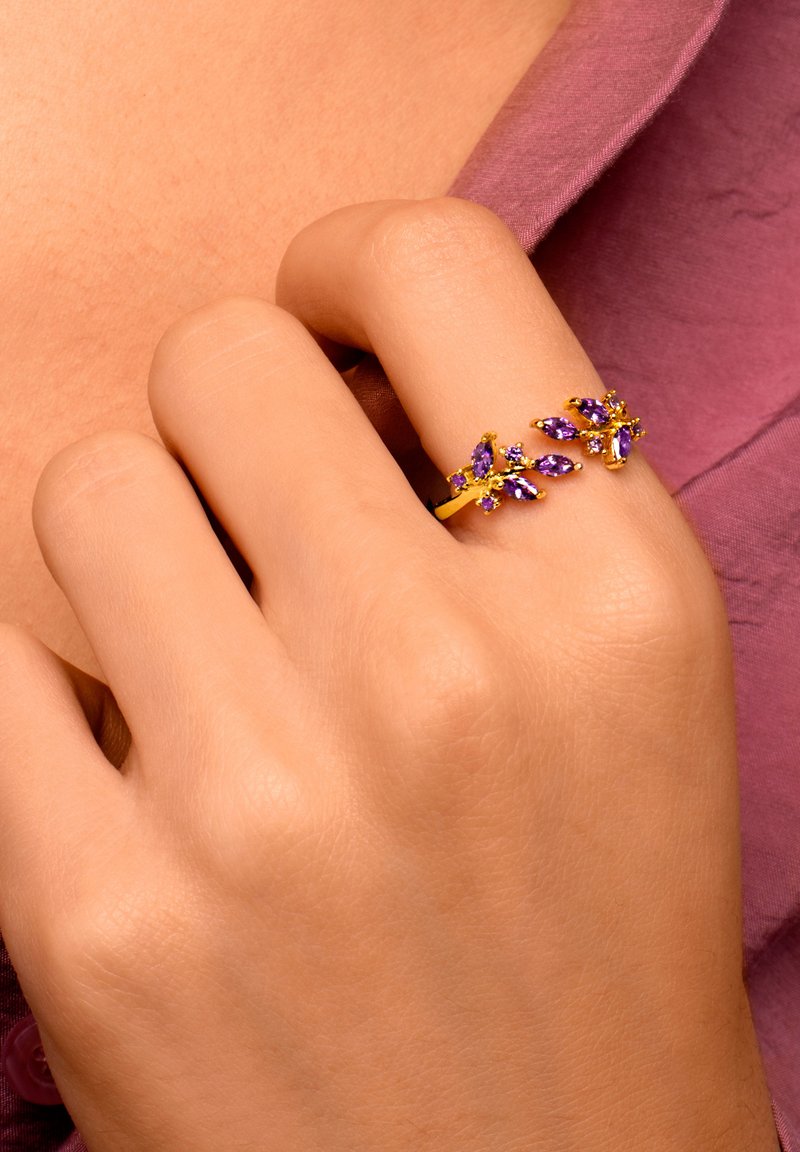 Anello d'oro con gemme viola disposte in un design botanico. La mano è mostrata appoggiata su uno sfondo di tessuto testurizzato.