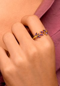Guldring med lila ädelstenar arrangerade i bladform, som ligger på en hand med en slät hudstruktur, mot en lila tygbakgrund.