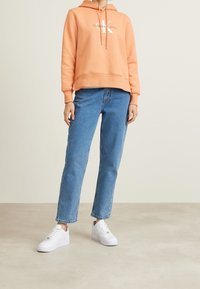 Coral hoodie with white logo, paired with relaxed-fit blue jeans and white sneakers. The fabric appears soft and comfortable.