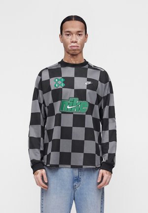 Persoană cu vitiligo purtând o bluză Nike neagră și gri în carouri cu mâneci lungi și blugi bleu deschis, stând pe un fundal alb simplu.