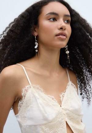 Jeune femme aux cheveux bouclés portant de longues boucles d'oreilles dorées avec des perles blanches et un caraco crème orné de dentelle.