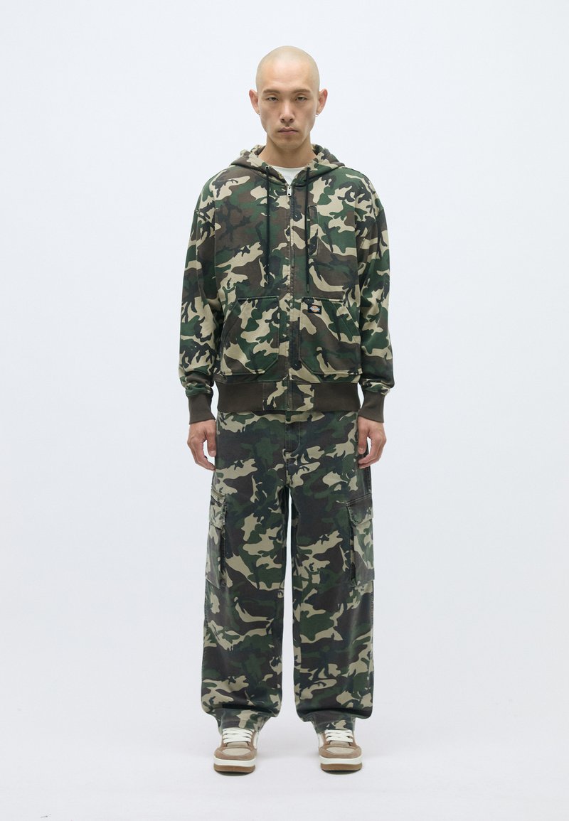 Maskējošs džemperis ar rāvējslēdzēju priekšā un kanguru kabatām, apvienots ar saskaņojošām platiem biksēm ar sānu kabatām.