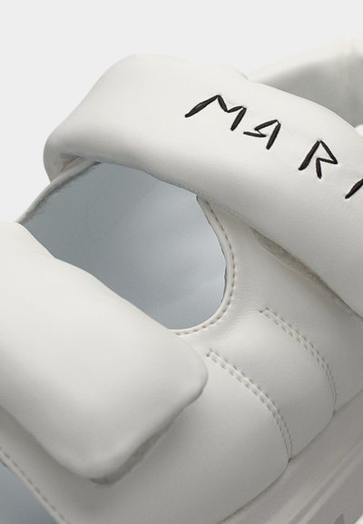 Λευκό δερμάτινο παπούτσι με στρογγυλεμένο, μινιμαλιστικό σχέδιο, που διαθέτει κομμένη περιοχή και ραμμένα διακοσμητικά, σημαδεμένο με μαύρα γράμματα.