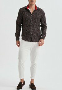 Chemise à motifs sombres avec des formes colorées, détail de col rouge ; associée à un pantalon blanc et des mocassins bruns.