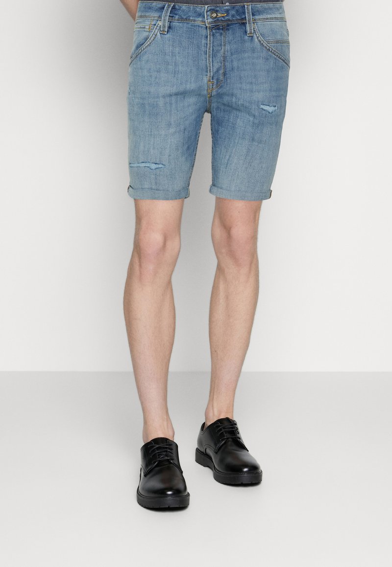 Pantalones cortos de denim en azul claro con un diseño desgastado; con puños enrollados, bolsillos estándar y acompañados de zapatos negros con cordones.