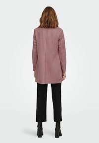 Langer pinker Wildledermantel mit geradem Schnitt, einer klaren hinteren Naht und langen Ärmeln, kombiniert mit schwarzer Hose und Stiefeletten mit Absatz.