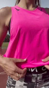 Woman adjusting bright pink Nike tank top, wearing black and white geometric leggings, standing on outdoor running track.