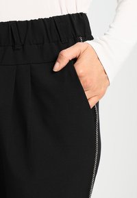 Pantalon noir avec une ceinture élastique, présentant une poche latérale et une bande d'accent blanche le long de la couture. Texture de tissu lisse.