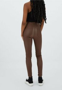 Bruine high-waisted skinny broek van een gladde, rekbare stof. Twee achterzakken, gecombineerd met zwarte sneakers. Achteraanzicht tegen een neutralere achtergrond.