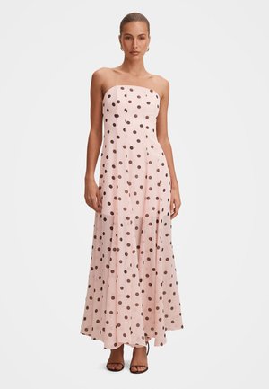 Femme portant une robe longue rose clair sans bretelles avec des pois noirs, associée à des talons ouverts noirs et des boucles d'oreilles en or en forme de créoles.