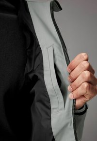 Interiorul jachetei gri deschis prezintă o textură netedă, căptușeală neagră și un buzunar lateral. O mână apucă ferm fermoarul pentru închidere.