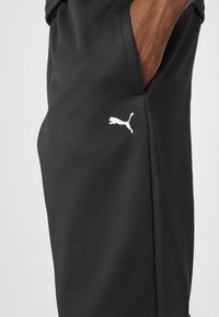 Svarta träningsbyxor i slät, texturerad tyg; har en vit Puma-logotyp på sidan; designade med sidofickor.