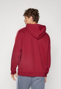 Red hooded sweatshirt with ribbed cuffs and hem, featuring white stripes along the shoulders, made of soft, textured fabric.