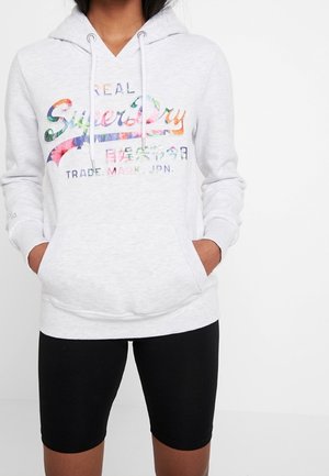 Witte hoodie met kleurrijk "Superdry" logo en Japanse tekst, gedragen door een persoon met handen in de voorzak, gecombineerd met zwarte shorts.