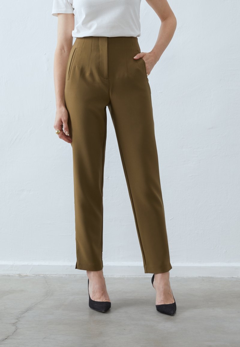 Femme portant un pantalon taille haute vert olive avec des fentes latérales, associé à des talons noirs à bouts pointus et un top blanc à manches courtes.