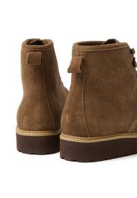 Stivaletti in suede marrone con tacco leggermente rialzato, due linguette e fori per lacci. Presentano una suola a contrasto con cuciture visibili.