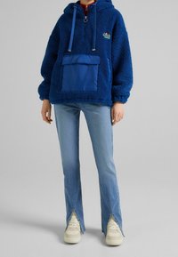 Blå fleecehuva med dragkedja vid halsen, framficka i en ljusare blå färg och snoddar. Matchat med ljusblå utsvängda jeans och sneakers.