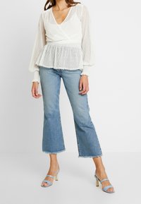 Blusa bianca con texture, maniche a palloncino trasparenti e vita a peplo, abbinata a jeans svasati azzurri e sandali a tacco azzurro cielo.