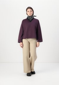 Felpa oversized viola con scollo a V, pantaloni beige a gamba larga e foulard nero. Ha maniche lunghe e una silhouette rilassata.